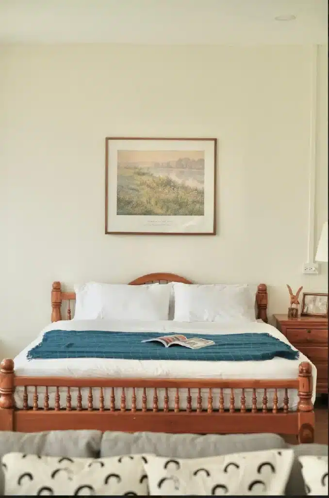 ที่พักฉะเชิงเทรา เตียงที่จัดอย่างประณีตพร้อมผ้าห่มสีน้ำเงินและหมอนสีขาว ภายใต้กรอบภาพวาดทิวทัศน์แบบชนบทบนผนังสีเบจของโรงแรมฉะเชิงเทรา