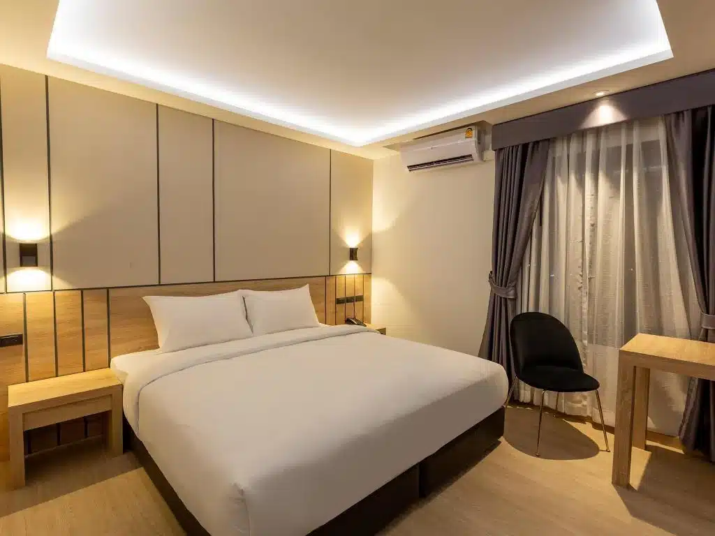 ห้องพักในโรงแรมทันสมัยมีเตียงขนาดใหญ่พร้อมผ้าปูที่นอนสีขาว หัวเตียงไม้ แสงไฟโดยรอบ โต๊ะทำงานพร้อมเก้าอี้ และผ้าม่านประดับใน โรงแรมฉะเชิงเทรา