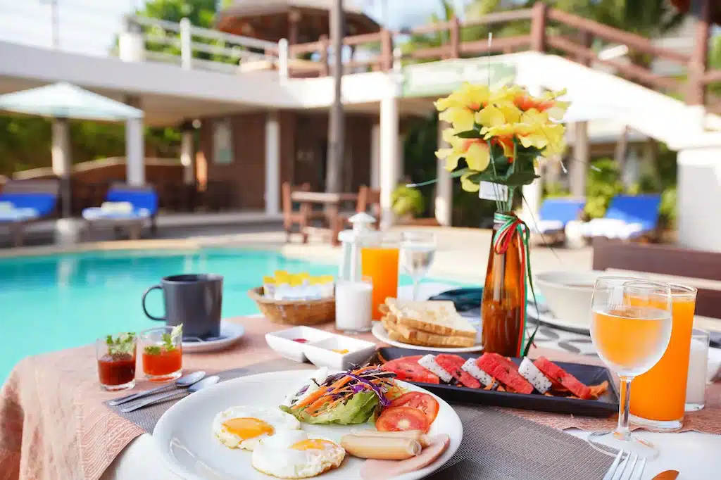 การตั้งค่าอาหารเช้ากลางแจ้งพร้อมวิวสระว่ายน้ำที่โรงแรมปราณบุรีติดทะเล มีไข่ ผลไม้ เครื่องดื่ม และการจัดดอกไม้บนโต๊ะ