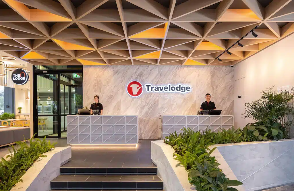 พนักงานต้อนรับสองคนยืนอยู่หลังโต๊ะในล็อบบี้ Travelodge อันทันสมัย พร้อมด้วยเพดานทรงเรขาคณิตและต้นไม้ในร่ม พร้อมที่จะช่วยเหลือผู้เข้าพักในการวางแผนการเดินทางสำหรับเที่ยวเอง