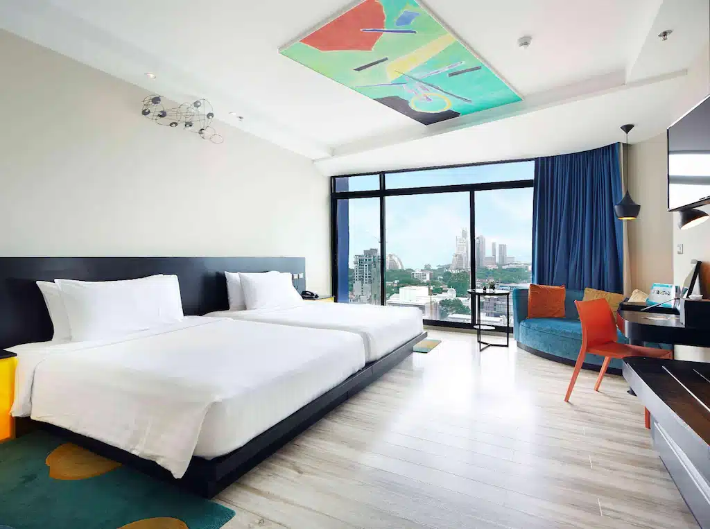 ห้องพักโรงแรมทันสมัยพร้อมเตียง 2 เตียง พรมปูพื้นสีสันสดใส และหน้าต่างบานใหญ่ที่มองเห็นเส้นขอบฟ้าของเมืองพัทยา ที่พักพัทยา