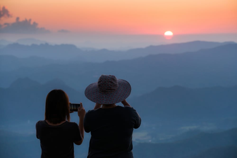 คน 2 คนหันหน้าออกจากกล้องเพื่อถ่ายรูปพระอาทิตย์ตกดินหลากสีสันเหนือทิวเขาด้วยโทรศัพท์ ที่พักอุ้มผาง