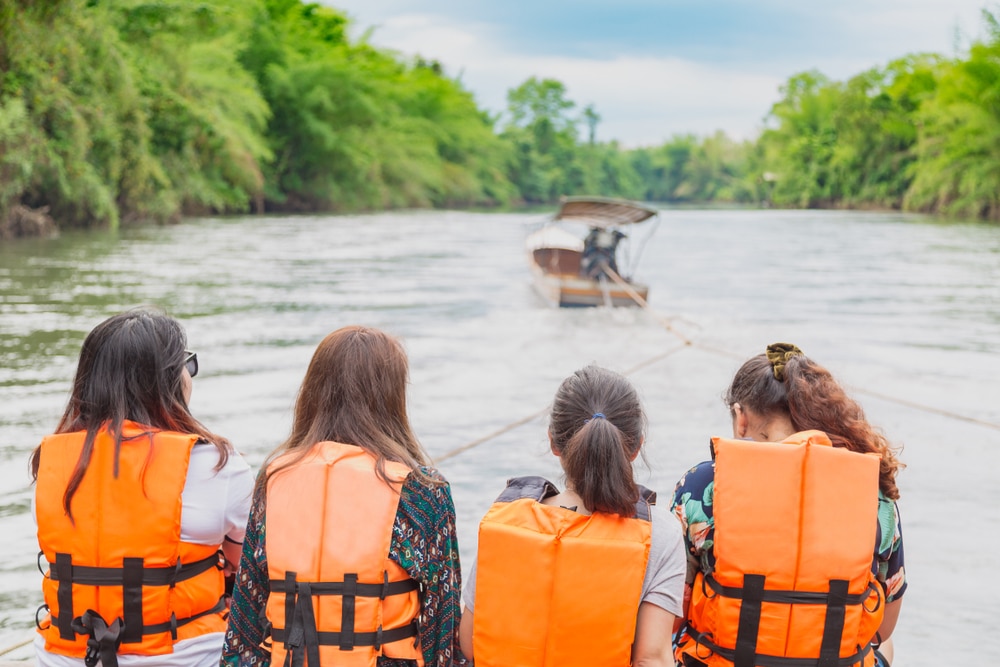 คนสี่คนสวมเสื้อชูชีพสีส้มนั่งบนเรือ โดยหันหน้าออกจากกล้อง ขณะที่เรือกำลังถูกลากไปตามแม่น้ำที่รายล้อมไปด้วยต้นไม้เขียวขจี ช่วงเวลาอันเงียบสงบนี้รวบรวมแก่นแท้ของการท่องเที่ยวกาญจนบุรี ไว้เป็นภาพที่สมบูรณ์แบบสำหรับใครก็ตามที่กำลังวางแผนเที่ยวกาญจนบุรี 1 วัน ท่องเที่ยวกาญจนบุรี