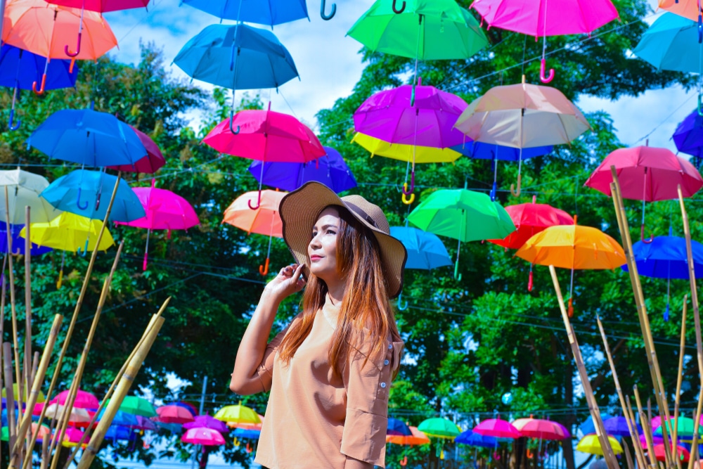 ผู้หญิงสวมหมวกกันแดดยืนอยู่ใต้ร่มสีสันสดใสที่แขวนไว้ในวันที่อากาศสดใสในนครนายกที่เที่ยว โดยมีต้นไม้ขึ้นเป็นฉากหลัง