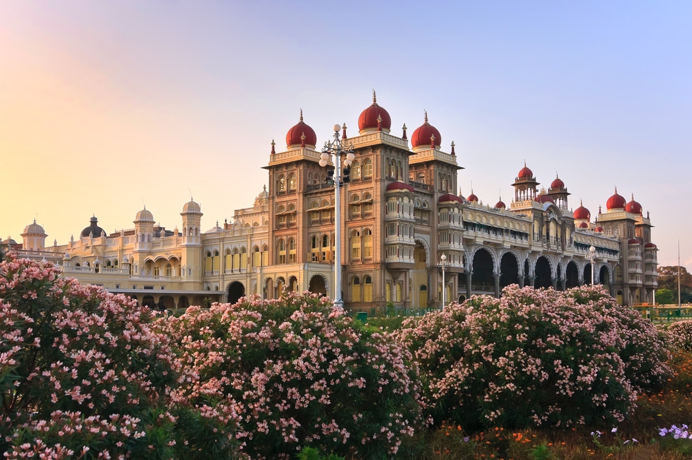 พระราชวังไมซอร์ในอินเดีย อาคารประวัติศาสตร์อันยิ่งใหญ่ที่มีโดมสีแดง สว่างไสวด้วยแสงพระอาทิตย์ตกสีทอง ล้อมรอบด้วยสวนที่บานสะพรั่งสีชมพูและสีขาวอันเขียวชอุ่ม