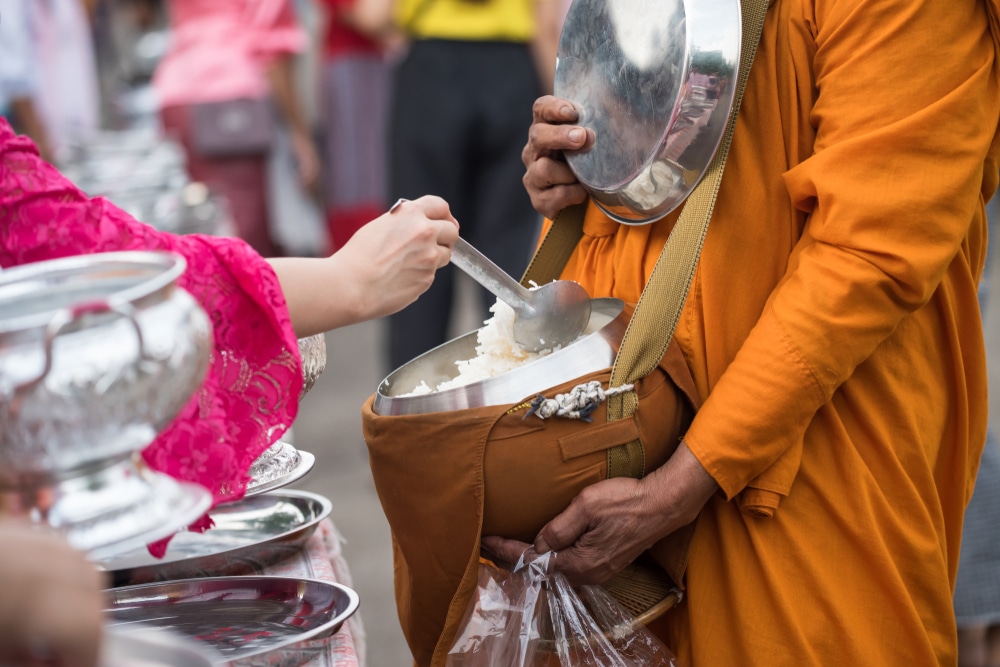 ในระหว่างการเยือนกาญจนบุรี บุคคลที่สวมเสื้อคลุมสีชมพูกำลังตักอาหารลงในชามที่พระภิกษุในชุดสีส้มถือในระหว่างพิธีกรรมหรือพิธีการ