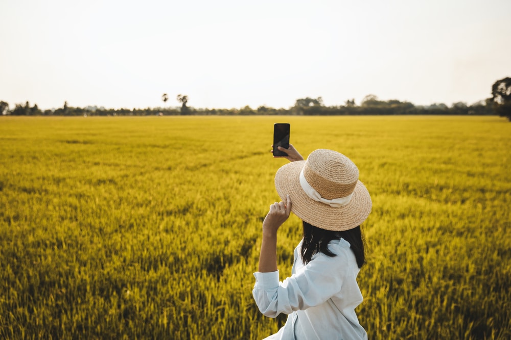 คนสวมหมวกฟางถ่ายรูปด้วยสมาร์ทโฟนในทุ่งหญ้าเขียวขจีอันกว้างใหญ่ในวันที่อากาศแจ่มใส เก็บภาพความงดงามของเที่ยวนครนายก