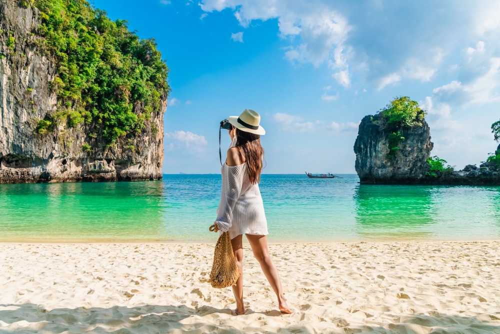 ผู้หญิงสวมหมวกและเดรสเดินไปตามชายหาดเขตร้อนบนเกาะพีพี โดยมีน้ำทะเลสีฟ้าใสและมีหินขนาดใหญ่เป็นฉากหลัง เกาะห้อง