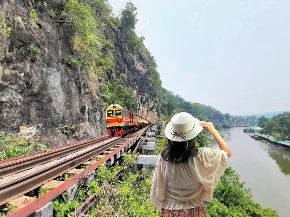 คนสวมหมวกสีขาวหันหน้าหนี ท่องเที่ยวกาญจนบุรี มองเห็นรถไฟที่วิ่งบนรางเลียบหน้าผาและแม่น้ำ จับภาพความงามอันเงียบสงบของเที่ยวกาญจนบุรี 1 วัน
