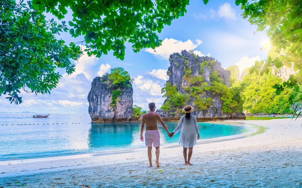 คู่รักเดินจูงมือกันบนหาดทรายมุ่งหน้าสู่ทะเลที่เกาะพีพี โดยมีโขดหินโผล่อยู่เบื้องหลังภายใต้ท้องฟ้าแจ่มใส เกาะห้อง