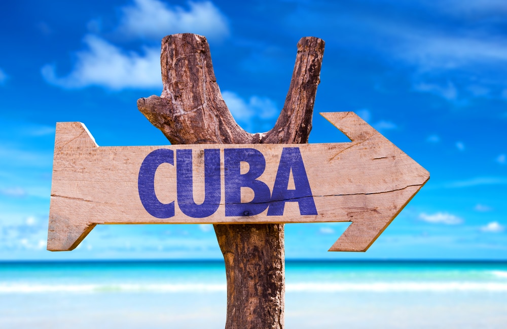 ป้ายลูกศรไม้ที่มีคำว่า CUBA ชี้ไปทางขวา ตั้งอยู่บนลำต้นของต้นไม้ โดยมีชายหาดและท้องฟ้าสีครามเป็นฉากหลัง ชวนให้นึกถึงการหลบหนีไปยังทะเลในทะเลลึกในโลก ทะเลที่สวยที่สุดในโลก