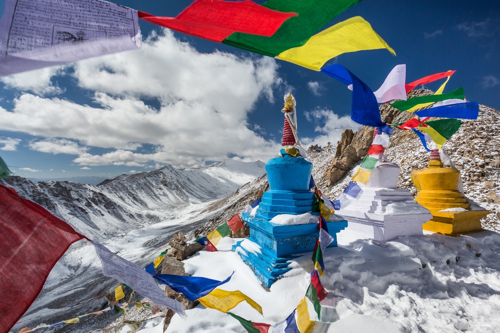ธงสวดมนต์สีสันสดใสและสถูปพุทธบนเส้นทางภูเขาหิมะในอินเดียภายใต้ท้องฟ้าสีฟ้าสดใสพร้อมเมฆ