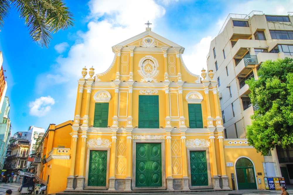 โบสถ์โคโลเนียลสีเหลืองและสีขาวที่มีประตูสีเขียวขนาบข้างด้วยอาคารสมัยใหม่ภายใต้ท้องฟ้าสีครามในมาเก๊า เที่ยวมาเก๊า
