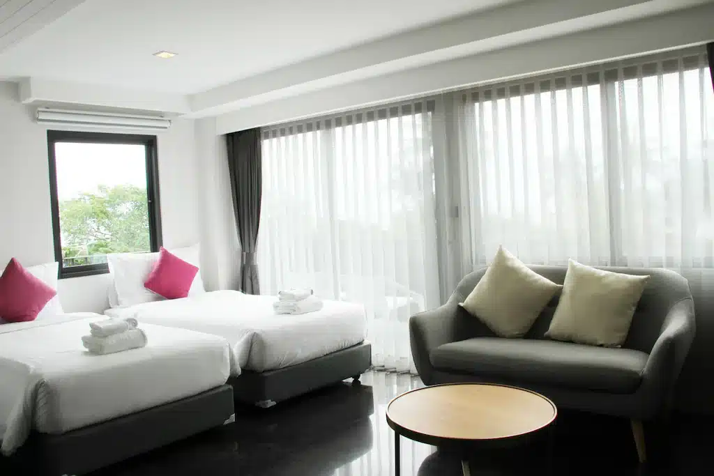ห้องพักในโรงแรมที่สว่างสดใสพร้อมเตียงแฝด 2 เตียง ชุดผ้าปูเตียงสีขาว และหมอนสีม่วง มีโซฟาสีเทาพร้อมเบาะรองนั่งสีซีดและโต๊ะกลมเล็กๆ หน้าต่างสูงจ ที่พักริมน้ำใกล้กรุงเทพ ากพื้นจรดเพดานมีผ้าม่านโปร่ง มองเห็นวิวสภาพแวดล้อมอันเงียบสงบที่ริมน้ำใกล้กรุงเทพแห่งนี้