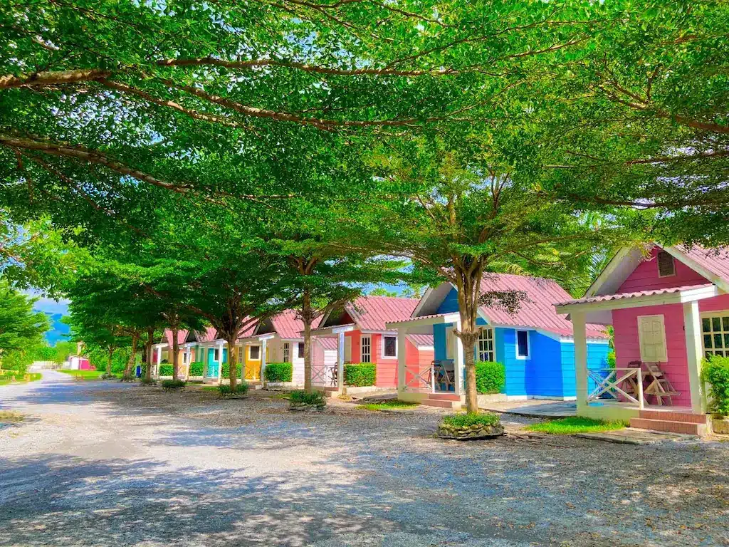 บ้านหลังเล็กๆ หลากสีสันที่มีหลังคาสีเขียว ล้อมรอบด้วยต้นไม้สีเขียวชอุ่มในวันที่อากาศแจ่มใส เป็นสถานที่พักผ่อนที่งดงาม ควบคุมริมน้ำใกล้กทม เหล่านี้มีการทาสีด้วยสีสดใส เช่น สีเหลือง สีฟ้า สีชมพู และสีม่วง ที่พักริมน้ำใกล้กรุงเทพ