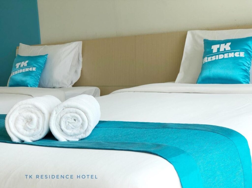 เตียงแฝดที่ตกแต่งอย่างประณีต 2 เตียงตกแต่งด้วยสีน้าน แต่ละเตียงตกแต่งด้วยหมอน "TK Residence" มีผ้าเช็ดตัวสีขาวม้วนอยู่บนเตียงหนึ่งเตียง เหมาะสำหรับการพักผ่อนหลังจากสำรวจสถานที่ท่องเที่ยวน่าน
