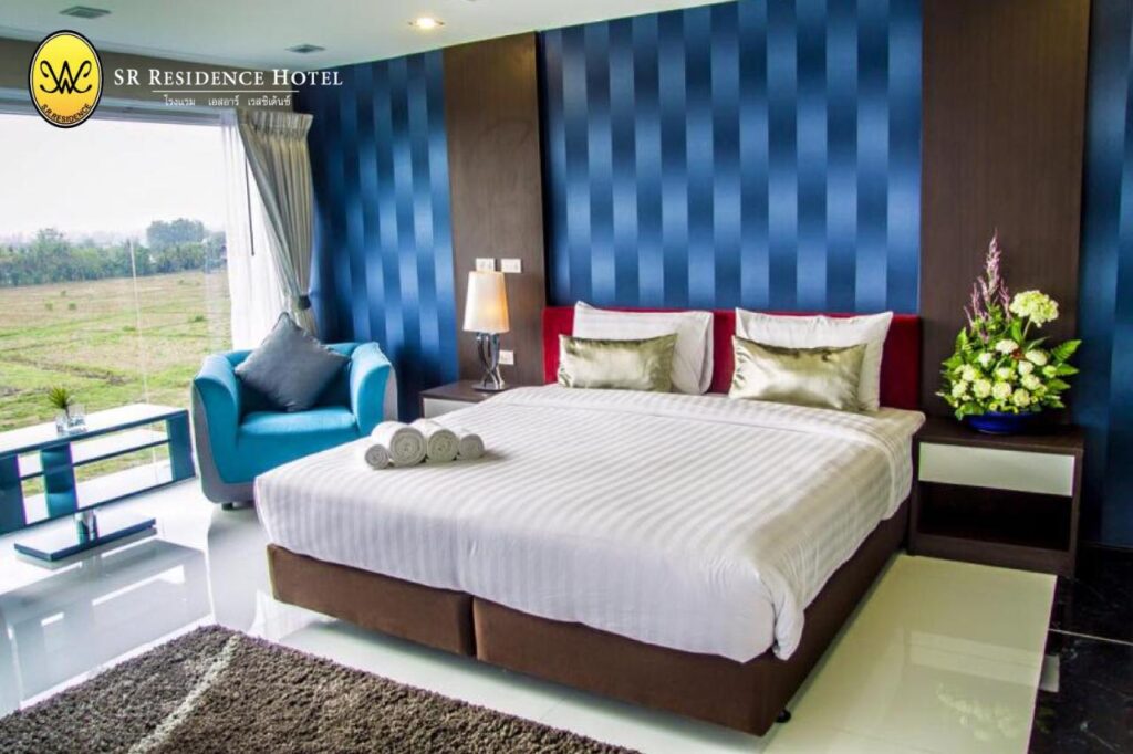 ห้องพักในโรงแรมทันสมัยพร้อมเตียงคู่ วอลล์เปเปอร์ลายทาง โต๊ะข้างเตียงพร้อมโคมไฟ อาร์มแชร์สีน้ำเงินข้างหน้าต่างบานใหญ่ และผ้าเช็ดตัวใหม่บนเตียง โลโก้ที่มุมซ้ายบนเขียนว่า "SR Residence Hotel" ที่พักใกล้วัดผาซ่อนแก้ว