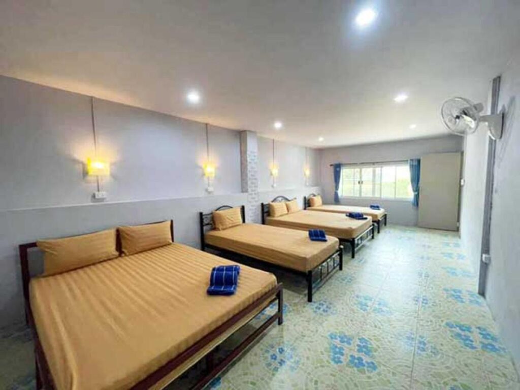 ห้องพักกว้างขวางพร้อมเตียงคู่ 4 เตียง แต่ละเตียงมีผ้าคลุมเตียงสีน้ำตาลและผ้าเช็ดตัวสีฟ้า ภายใต้แสงไฟสีเหลืองอันอบอุ่น ห้องพักมีพื้นกระเบื้อง หน้าต่างบานใหญ่ และพัดลมติดผนัง เหมาะสำหรับผู้ที่ต้องการความสะดวกสบายกาฬสินธุ์