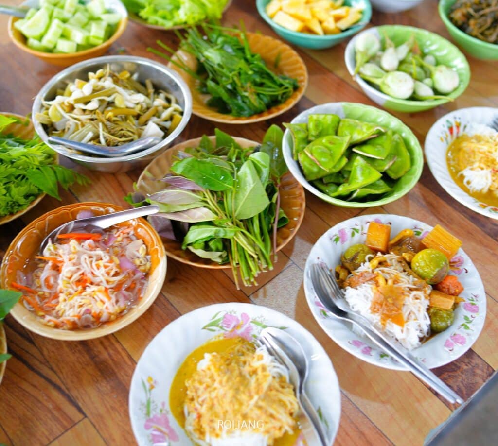 ร้านอาหารพังงา อาหารหลากสีสันและผักสดหลากหลายชนิด เช่น บะหมี่ ผักใบเขียว สมุนไพร และแตงกวาหั่นบาง ๆ จัดวางอยู่บนโต๊ะไม้ที่ร้านอาหารเมืองพังงา