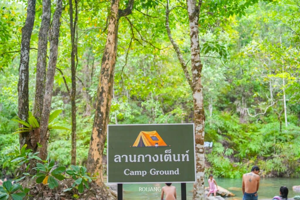 ป้ายเขียนว่า "Camp Ground" ในพื้นที่ป่า โดยมองเห็นคนหลายคนอยู่เบื้องหลังใกล้แหล่งน้ำ เน้นที่เที่ยวตะกั่วป่าอันมีเสน่ห์แห่งหนึ่ง ที่เที่ยวตะกั่วป่า