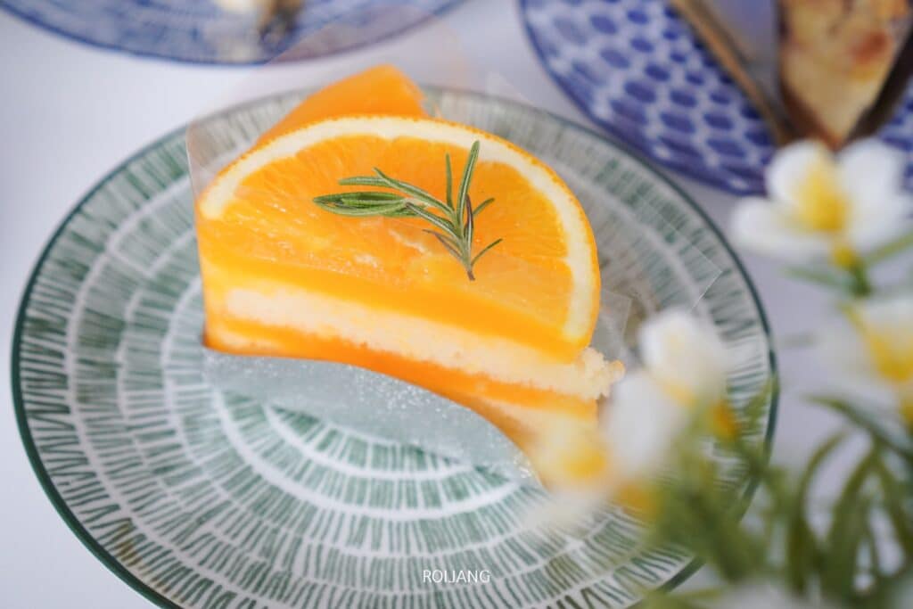 เค้กส้มชิ้นหนึ่ง ตกแต่งด้วยชิ้นส้มและโรสแมรี่ วางอยู่บนจานที่มีลวดลายสีเขียว เหมาะสำหรับรับประทานที่ร้าน คาเฟ่ตะกั่วป่า