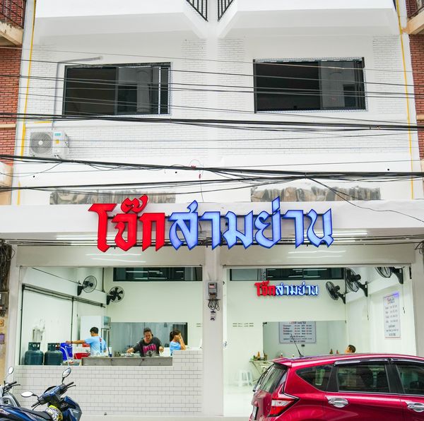 มุมมองถนนของร้านอาหาร ชาบูคอง ภูเก็ต มีป้ายเป็นภาษาไทย ตัวอาคารมีส่วนหน้าอาคารสีขาวและพื้นที่รับประทานอาหารด้านหน้ากระจก ข้างในมีคนไม่กี่คนและรถจอดอยู่ข้างหน้า ตอกย้ำถึงแก่นแท้ของ ร้านอาหารภูเก็ต