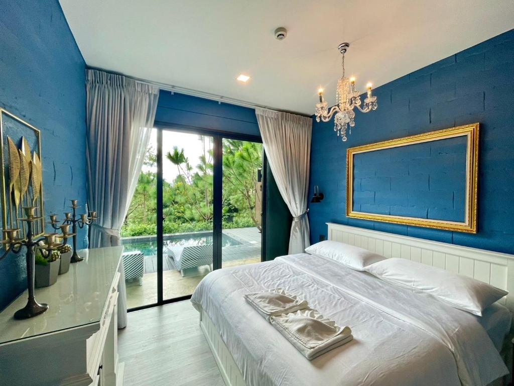 ห้องนอนที่มีผนังสีฟ้ามีเตียงสีขาว กระจกกรอบทอง และโคมระย้า ประตูกระจกเปิดออกสู่ลานเฉลียงพร้อมสระว่ายน้ำและแมกไม้เขียวขจีด้านนอก ที่พักใกล้วัดผาซ่อนแก้ว