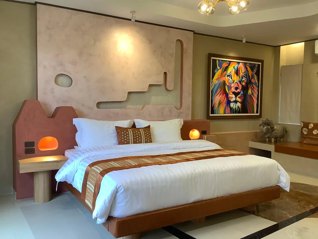 ห้องนอนทันสมัยมีเตียงคู่พร้อมชุดผ้าปูเตียงสีขาวและหัวเตียงไม้ที่ส่องสว่างด้วยโคมไฟข้างเตียง ภาพวาดสิงโตสีสันสดใสแขวนอยู่บนผนัง ที่พักใกล้วัดผาซ่อนแก้ว