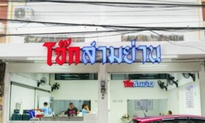 มุมมองภายนอกของร้านอาหารที่มีป้ายเขียนด้วยอักษรไทย แสดงให้เห็นคนงานภายในร้านกำลังยุ่งอยู่กับการเตรียมการ รถสีแดงจอดอยู่หน้าอาคาร บ่งบอกถึงบรรยากาศคึกคักของโจ๊กสามย่าน