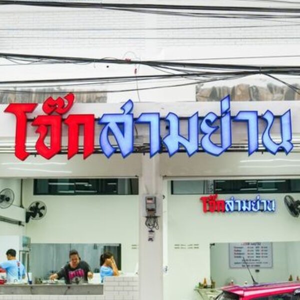 มุมมองภายนอกของร้านอาหารที่มีป้ายเขียนด้วยอักษรไทย แสดงให้เห็นคนงานภายในร้านกำลังยุ่งอยู่กับการเตรียมการ รถสีแดงจอดอยู่หน้าอาคาร บ่งบอกถึงบรรยากาศคึกคักของโจ๊กสามย่าน