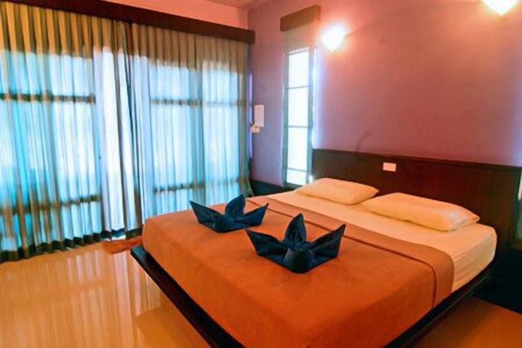ห้องนอนที่มีเตียงคู่ หงส์ผ้าเช็ดตัวสีน้ำเงินสองตัวบนเตียง ผนังสีม่วง และผ้าม่านหน้าต่าง ท่องเที่ยวเกาะลันตา