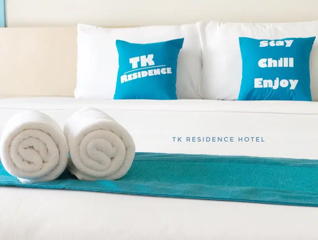 เตียงที่จัดอย่างประณีตพร้อมผ้าเช็ดตัวสีขาวม้วนอยู่บนรันเนอร์สีน้ำเงิน เตียงมีหมอนสีฟ้าสองใบที่มีป้ายกำกับว่า "TK Residence" และ "Stay Chill Enjoy" คำว่า "TK Residence Hotel" อยู่ในตำแหน่งรองชนะเลิศ เหมาะสำหรับผู้ที่ต้องการสำรวจสถานที่ท่องเที่ยวน่าน