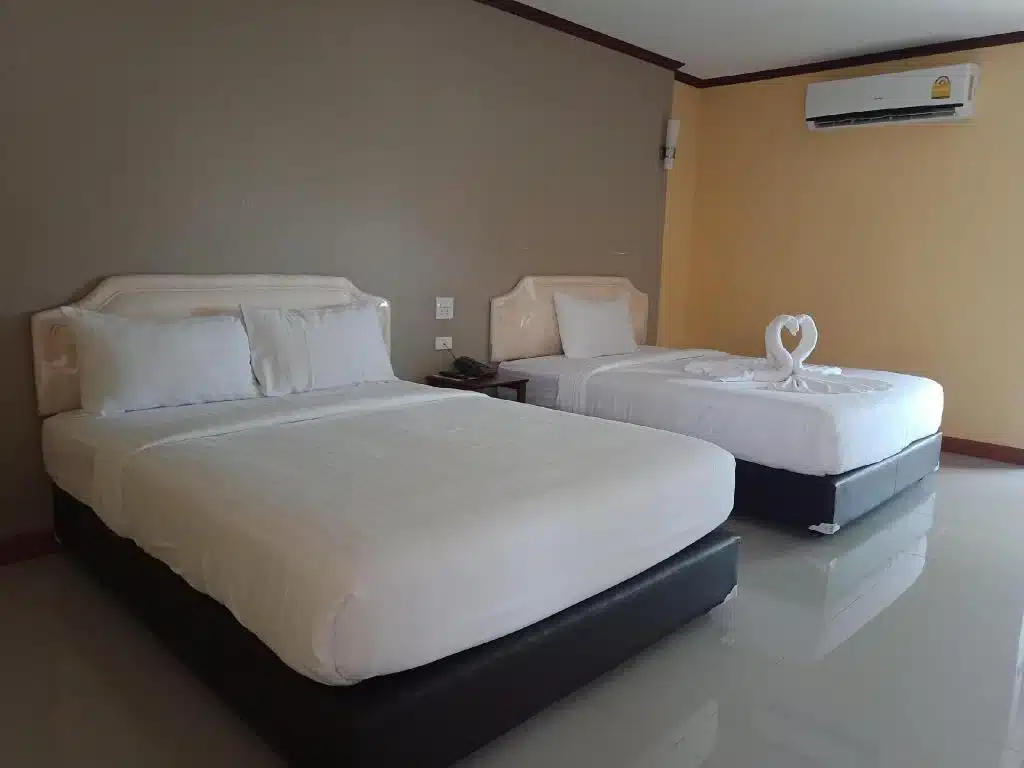 ห้องพักในโรงแรมที่มีเตียงที่จัดอย่างประณีต 2 เตียงพร้อมผ้าปูเตียงสีขาว ผ้าขนหนูพับเป็นรูปหงส์วางอยู่บนเตียงหนึ่งเตียง มีเครื่องปรับอากาศบนผนังและมีโต๊ะข้างเตียงระหว่างเตียง เพื่อให้แขกได้พักผ่อนอย่างสะดวกสบายในกาฬสินธุ์