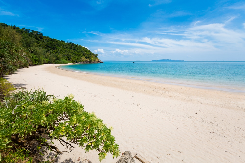 หาดทรายอันเงียบสงบพร้อมน้ำทะเลสีฟ้าครามใส ล้อมรอบด้วยพันธุ์ไม้สีเขียวชอุ่มภายใต้ท้องฟ้าสีครามสดใสและมีเมฆกระจายอยู่