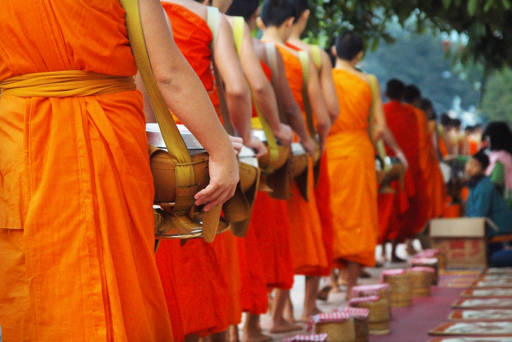 พระภิกษุในชุดสีส้มสดใสเดินเป็นขบวน แต่ละคนถือชาม มีตะกร้าวางอยู่บนพื้นข้างทาง เป็นภาพที่สวยงามตามแบบฉบับของหลวงพระบาง หลวงพระบาง