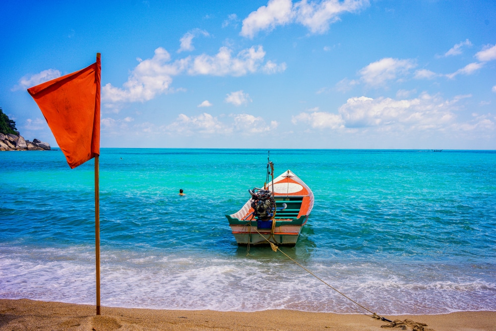 เรือลำเล็กทอดสมออยู่บนชายหาดเขตร้อนที่มีน้ำทะเลสีฟ้าใสและมีธงสีส้มอยู่เบื้องหน้า สถานที่ท่องเที่ยวสุราษ 