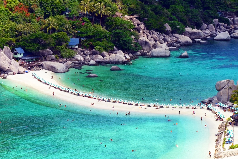 หาดทรายเล็กๆ โค้งระหว่างเกาะสองแห่งที่มีป่าไม้ น้ำสีฟ้าครามล้อมรอบชายหาด หลายๆ คนกำลังอาบแดดและว่ายน้ำ โดยมีร่มชายหาดเรียงรายอยู่ริมชายฝั่ง สถานที่ท่องเที่ยวสุราษ 