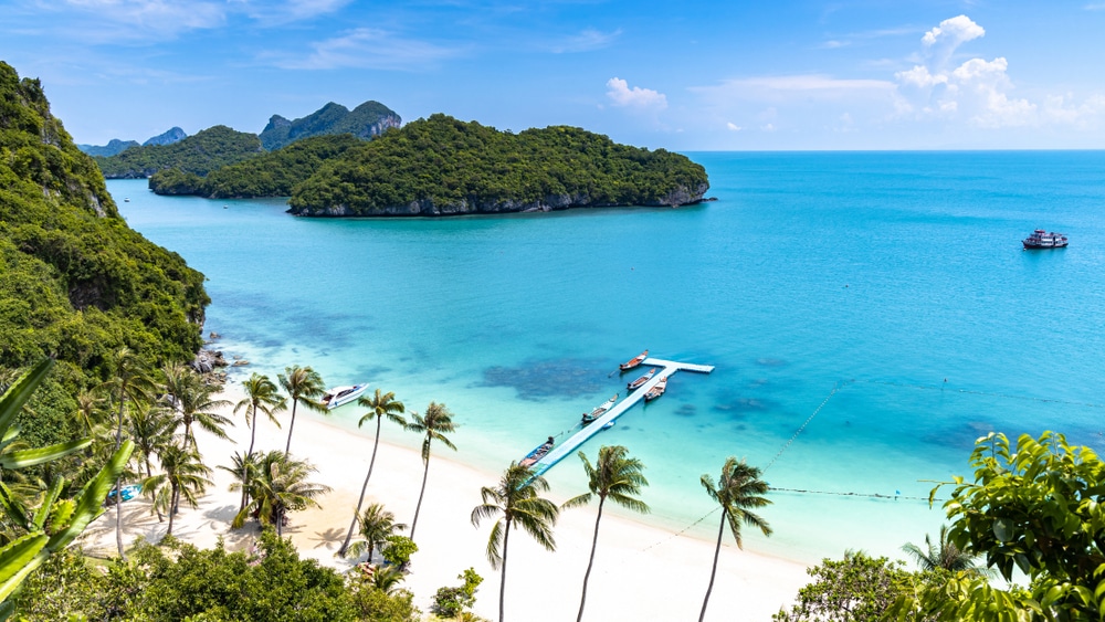 ฉากชายหาดอันเงียบสงบที่มีน้ำทะเลสีฟ้าครามใส หาดทรายขาว ท่าเรือที่มีเรือ ต้นปาล์ม และเกาะอันเขียวชอุ่มภายใต้ท้องฟ้าสีครามสดใส สถานที่ท่องเที่ยวสุราษ 