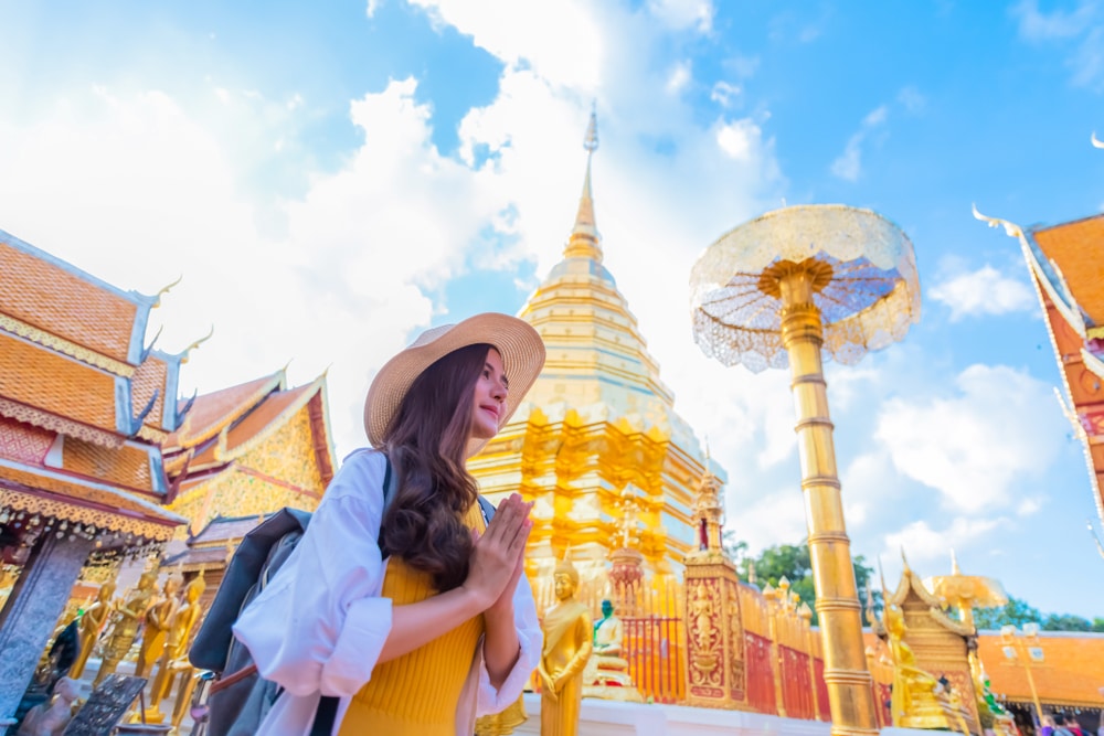 คนสวมหมวกและชุดสีเหลืองยืนประนมมือประสานกันในท่าสวดมนต์ ณ วัดสีทองใต้ท้องฟ้าสีครามสดใส ถ่ายภาพความงามอันเงียบสงบของสถาน ที่ท่องเที่ยวในเชียงใหม่