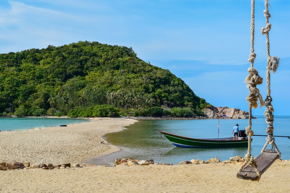 เรือลำหนึ่งจอดทอดสมออยู่ใกล้หาดทราย โดยมีเนินเขาสีเขียวชอุ่มเป็นฉากหลัง สถานที่ท่องเที่ยวสุราษ ชิงช้าเชือกแขวนอยู่เบื้องหน้า