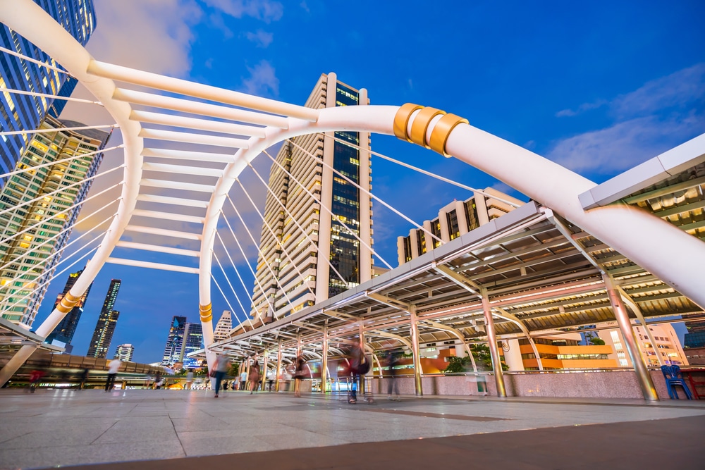 สะพานในเมืองสมัยใหม่ที่มีคานโค้งสีขาวและ ที่เดทในกรุงเทพกลางคืน ผู้คนเดินอยู่ข้างใต้ ล้อมรอบด้วยอาคารกระจกสูงในทิวทัศน์เมืองยามพลบค่ำ ทำให้เป็นสถานที่ที่เหมาะสำหรับการเดทในกรุงเทพตอนกลางคืน