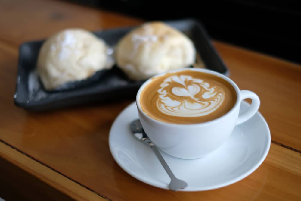 ถ้วยกาแฟที่มีงานศิลปะจากโฟมอันประณีตวางอยู่บนโต๊ะไม้ข้างจานที่บรรจุขนมอบ 2 ชิ้น ที่เที่ยวเกาะลันตา