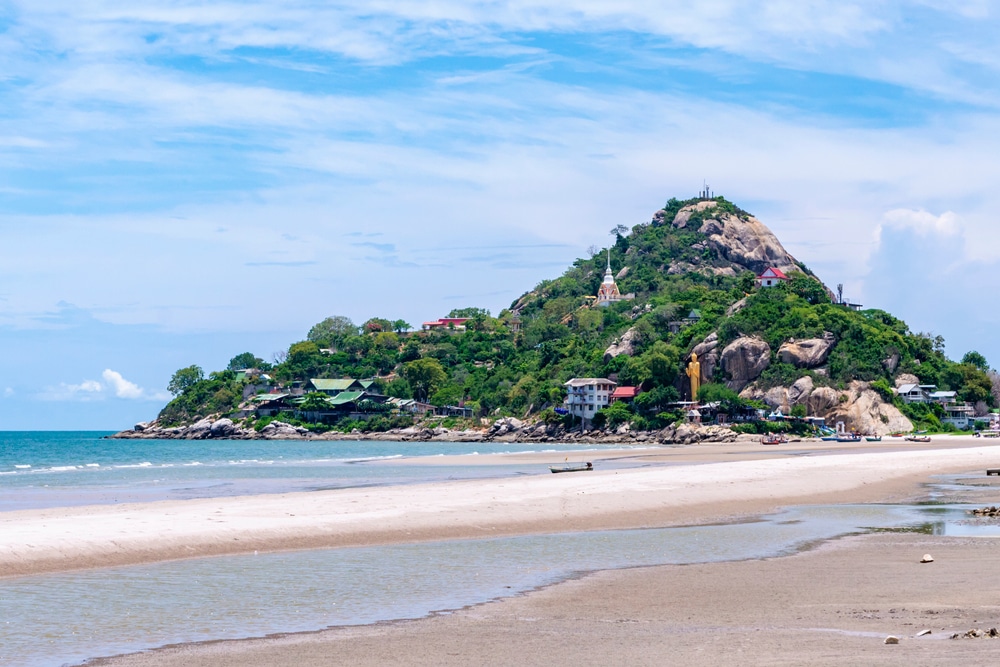 ชายหาดที่สวยงามพร้อมทิวทัศน์ของเนินเขาที่ปกคลุมไปด้วยความเขียวขจีและอาคารต่างๆ บรรจบกับมหาสมุทรสีฟ้าภายใต้ท้องฟ้ สถานที่ฮันนีมูนในไทย าที่มีเมฆบางส่วน ซึ่งเป็นสถานที่ที่สมบูรณ์แบบในฮอนีมูนในไทย