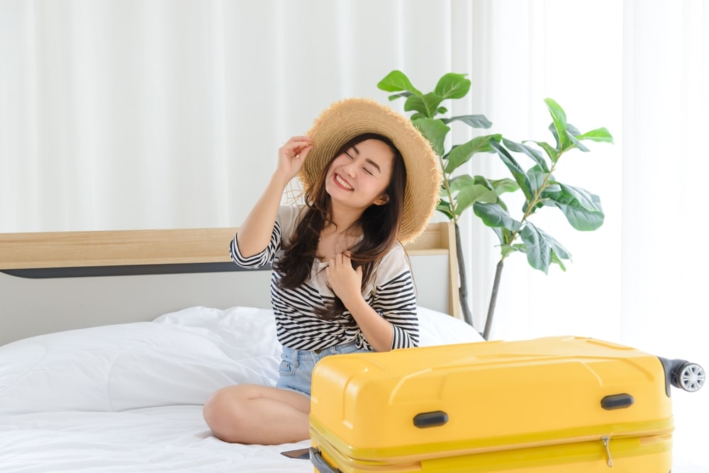 ผู้หญิงสวมหมวกฟางยิ้มกำลังนั่งอยู่บนเตียงข้างกระเป๋าเดินทางสีเหลือง โดยมีกระถางต้นไม้เป็นพื้นหลัง กำลังเตรียมตัวเข้าพักที่โรงแรมนี้ใกล้ผาซ่อนแก้ว