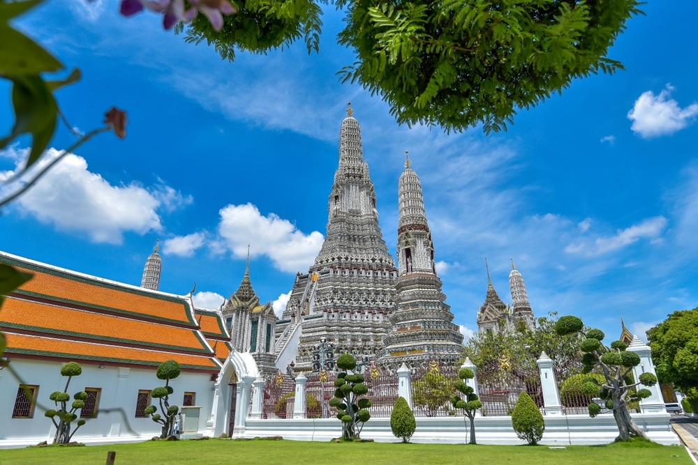 รูปภาพวัดอรุณ วัดพุทธในกรุงเทพฯ  ที่เดทในกรุงเทพกลางคืน ประเทศไทย โดดเด่นด้วยยอดแหลมสูงและสถาปัตยกรรมอันวิจิตรบรรจง ตัดกับท้องฟ้าสีครามและมีเมฆกระจัดกระจาย ต้นไม้ที่อยู่เบื้องหน้าทำให้ฉากดูน่าหลงใหล ณ วันที่ในกรุงเทพตอนกลางคืน