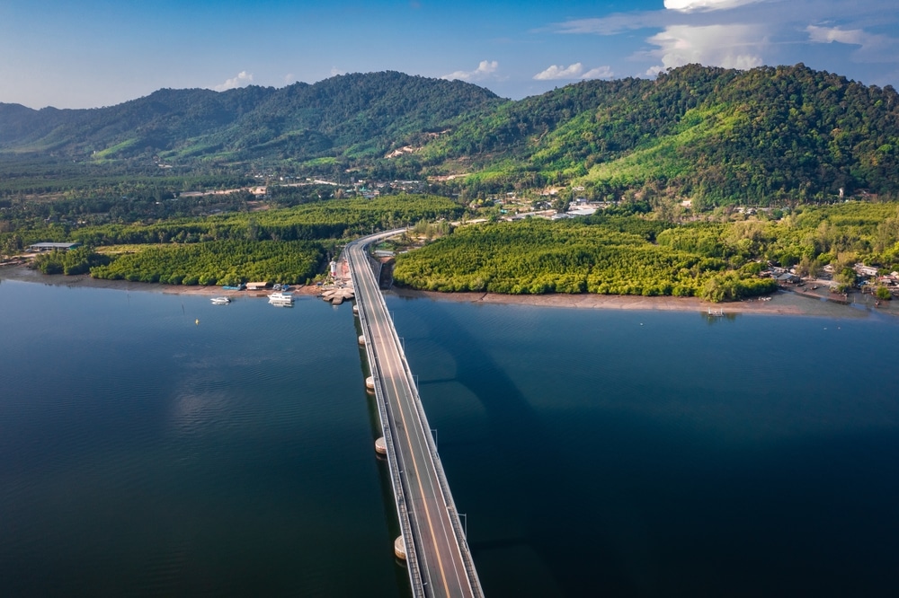 สะพานยาวทอดยาวเหนือแหล่งน้ำอันเงียบสงบ เชื่อมต่อกับภูมิทัศน์ภูเขาอันเขียวชอุ่มภายใต้ท้องฟ้าสีฟ้าใส ที่เที่ยวเกาะลันตา