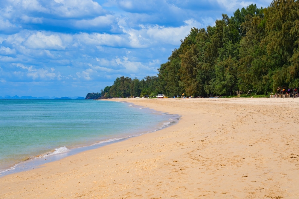 หาดทรายที่มีน้ำทะเลสีฟ้าอมเขียวอันเงียบสงบทางด้านซ้าย และมีแนวต้นไม้สูงทางด้านขวาใต้ท้องฟ้าที่มีเมฆบางส่วน ที่เที่ยวเกาะลันตา