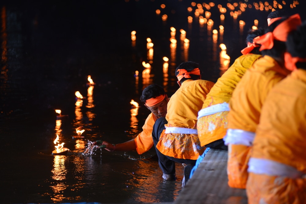 กลุ่มคนในชุดสีส้มจุดเทียนในแม่น้ำ โดยมีเปลวไฟเล็กๆ จำนวนมากสะท้อนอยู่ในน้ำในเวลากลางคืน เพื่อเฉลิมฉลอง งานลอยกระทง