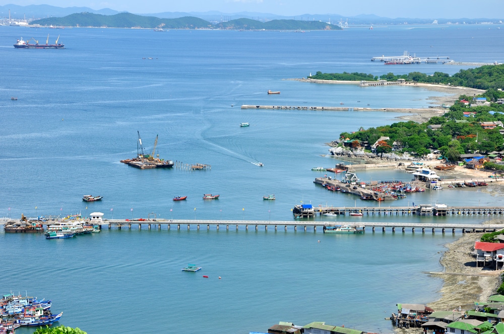 มุมมองทางอากาศของพื้นที่ชายฝั่งที่มีท่าเรือยาว เรือหลายลำ และอาคารต สถานที่ฮันนีมูนในไทย ามแนวชายฝั่ง เนินเขาและแนวชายฝั่งอันห่างไกลมองเห็นเป็นพื้นหลังภายใต้ท้องฟ้าสีฟ้าใส ทำให้เป็นสถานที่ที่เหมาะสำหรับพื้นที่ฮันนีมูนในไทย