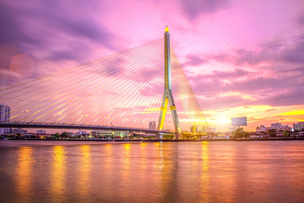 สะพานแขวนทอดข้ามแม่น้ำโดยมีทิวทัศน์เมืองเป็นพื้นหลังภายใต้ท้องฟ้ายาม ที่เดทในกรุงเทพกลางคืน พระอาทิตย์ตกดินที่เต็มไปด้วยสีสัน ทำให้เกิดบรรยากาศที่งดงามราวกับภาพวาดซึ่งอาจเป็นหนึ่งในสถานที่เดทที่ดีที่สุดในกรุงเทพกลางคืนได้อย่างง่ายดาย